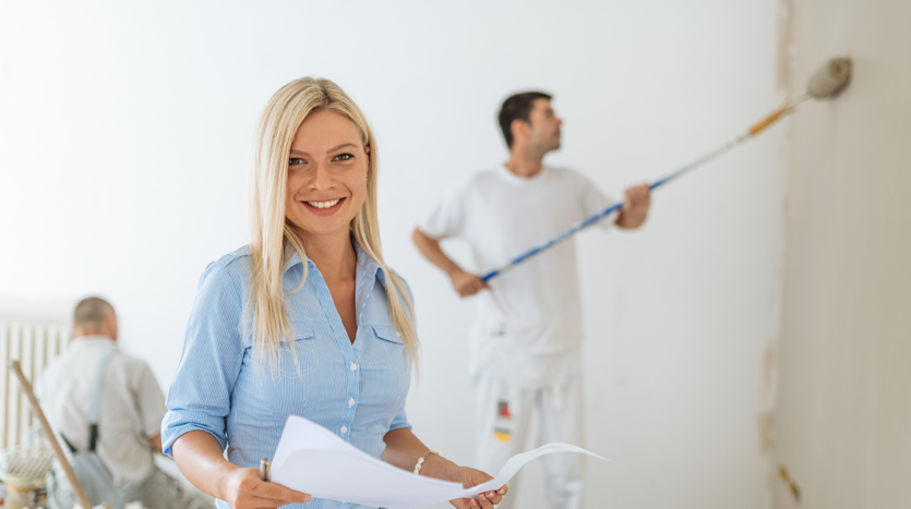 Smiling woman overseeing a home renovation project with workers painting the walls, highlighting property renovations in Phuket to increase rental appeal.